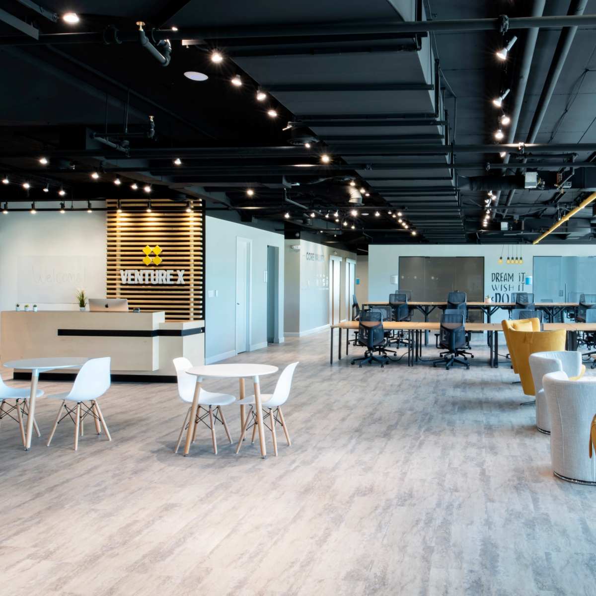 An open modern office space features a reception area, multiple seating arrangements, and a row of desks in a brightly lit environment.