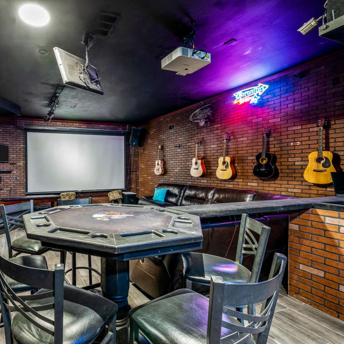 A game room features a poker table surrounded by chairs, a large projection screen, and wall-mounted guitars, all set against a brick backdrop.