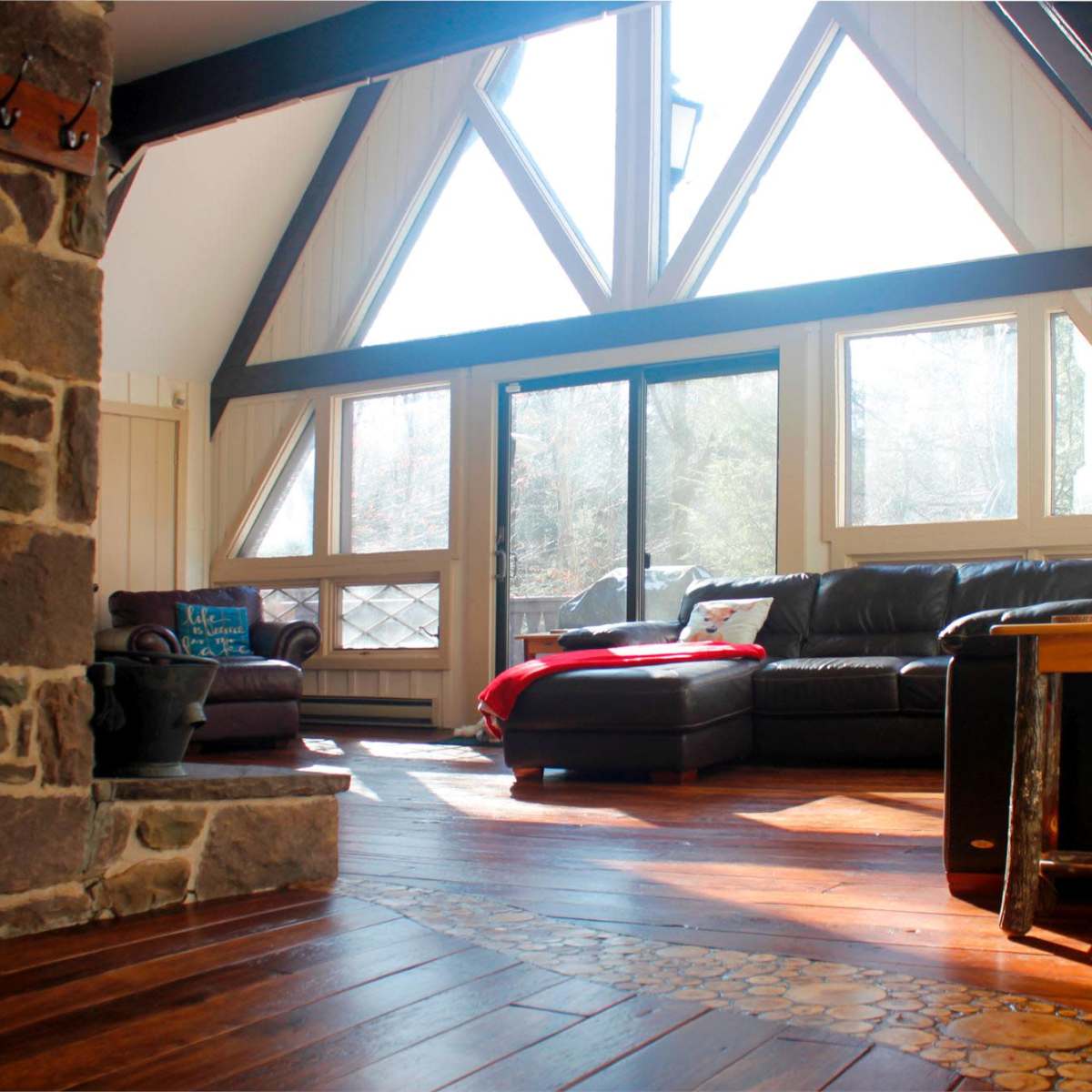 A cozy living room with large triangular windows, wooden floors, a stone fireplace, and a black leather sofa.