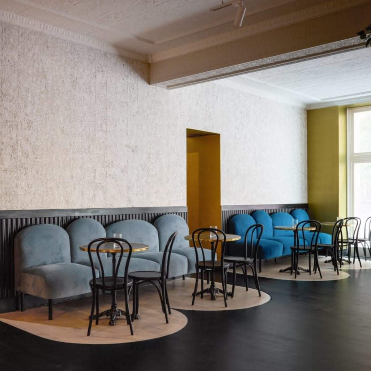 A modern café interior with blue and beige upholstered seating, round tables, and large windows allowing natural light to enter.