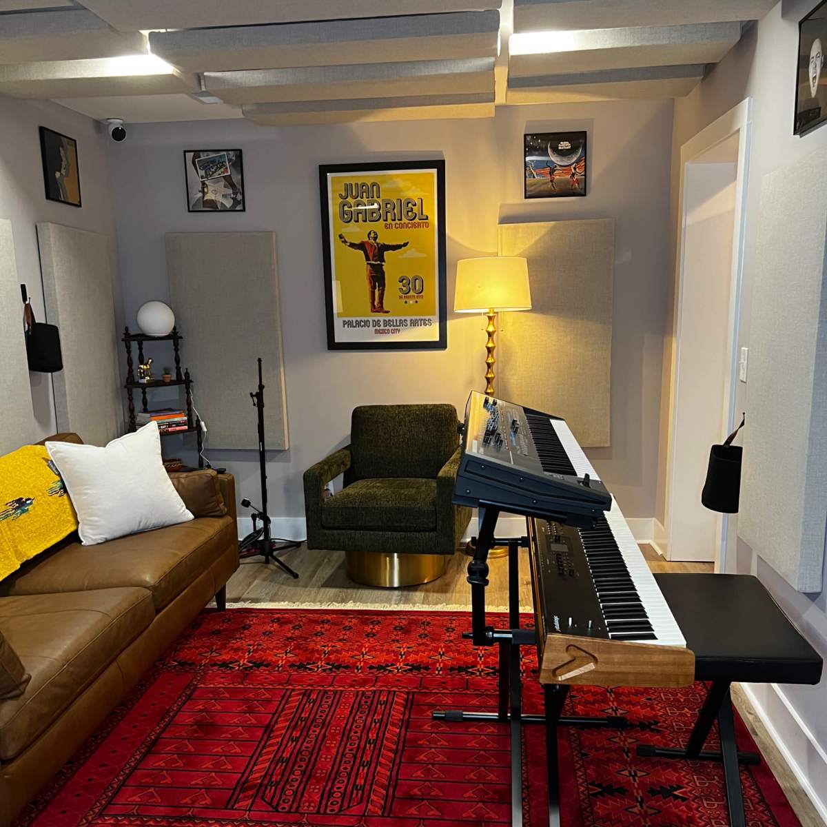A home studio with a brown couch, a keyboard on a stand, a round lamp, and framed artwork on the walls, all set on a red patterned rug.