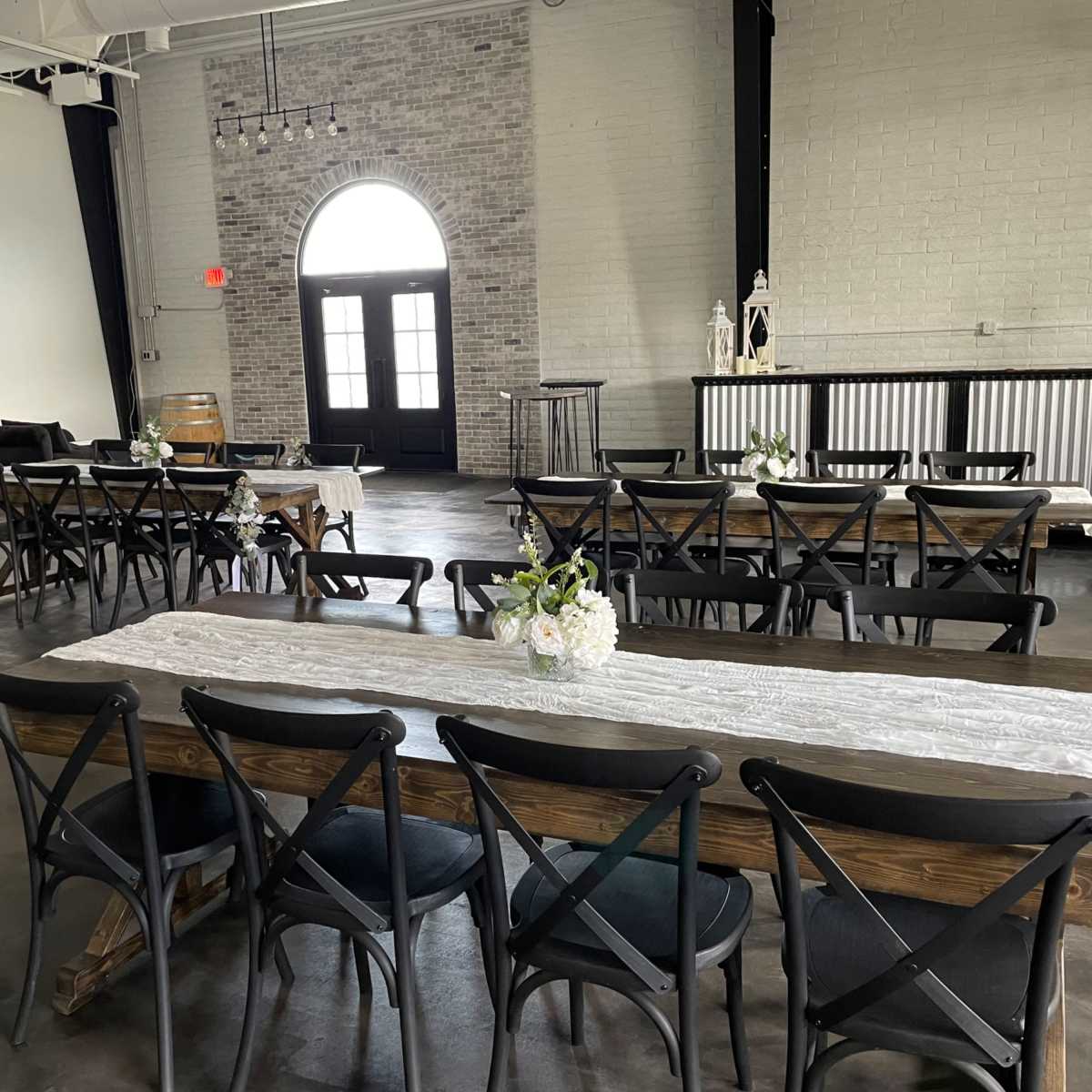 An indoor event space featuring several long wooden tables arranged with chairs and floral centerpieces, with large windows and a bar area in the background.