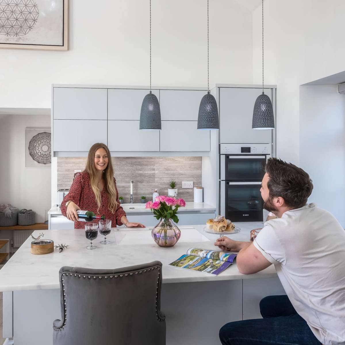 A woman serves drinks at a kitchen island while a man sits nearby, both in a modern kitchen setting.