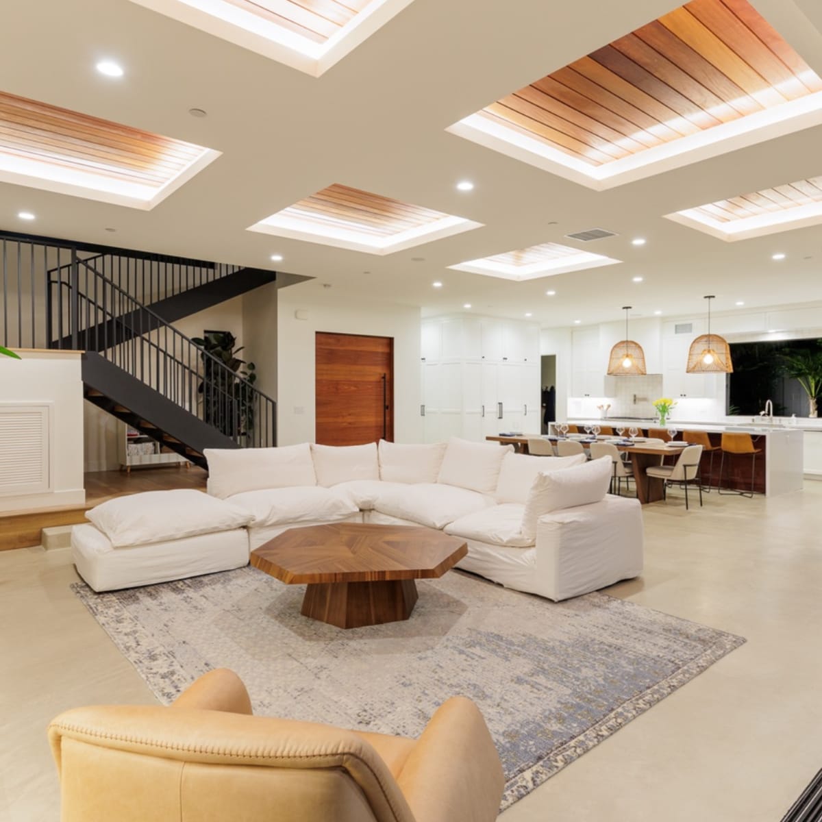 A modern living room with a white sectional sofa, a wooden coffee table, and a staircase leading to an upper level, adjacent to a dining area and kitchen.