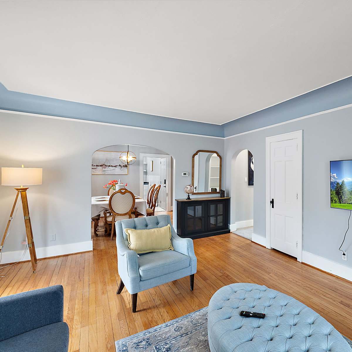 A living room with a light blue wall, a mounted television displaying a landscape, a light blue armchair, an ottoman, and a view into a dining area with a table and chairs.