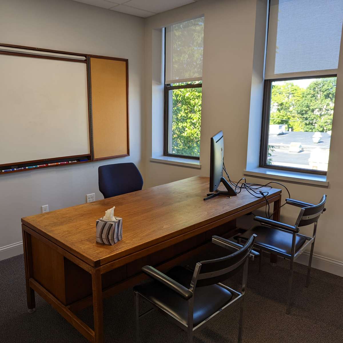 A small, well-lit meeting room features a wooden table, a chair, a computer monitor, and a whiteboard.