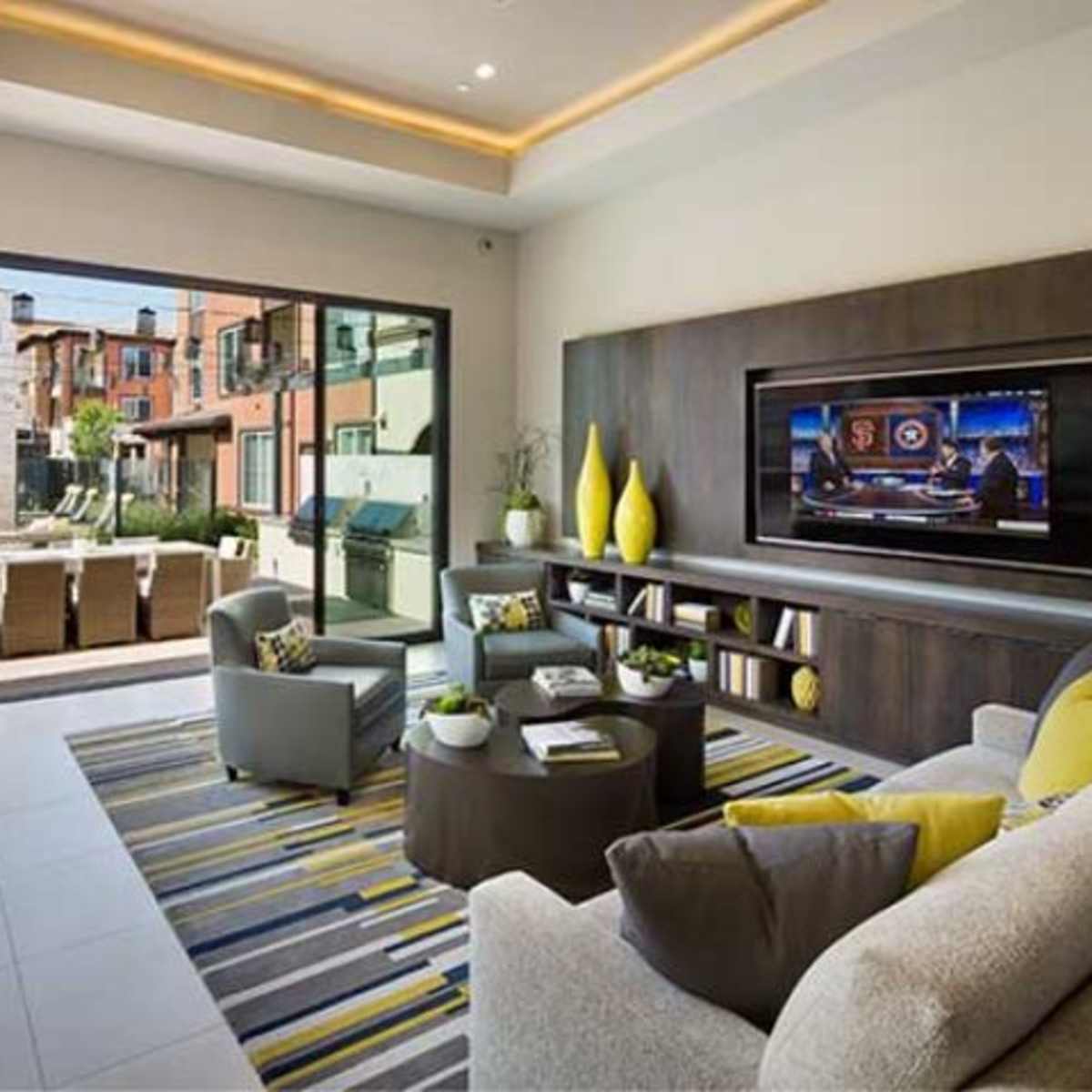 A modern living room features a large window showcasing an outdoor seating area, with a television mounted on the wall and a coffee table surrounded by gray chairs and a sofa.