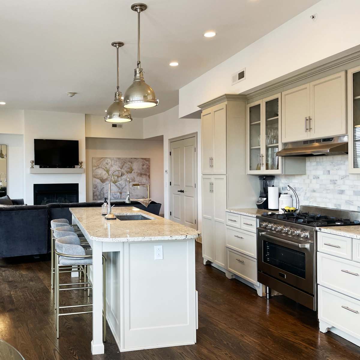 A modern kitchen with an island, featuring stainless steel appliances, a stone countertop, and a living area with a television in the background.