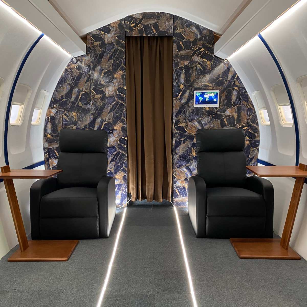 The interior of a private jet showcases two black reclining chairs flanking a central aisle, with a patterned wall and soft lighting.