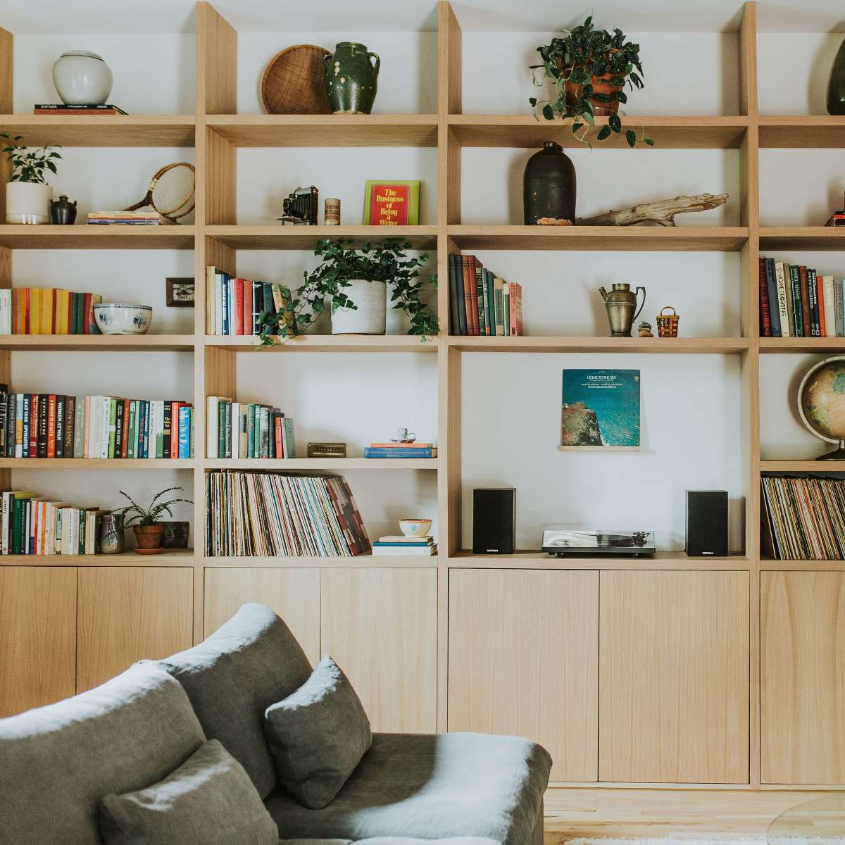 A wooden bookshelf filled with various books, plants, and decor items stands against a wall in a cozy living room, accompanied by a gray sofa and a coffee table.