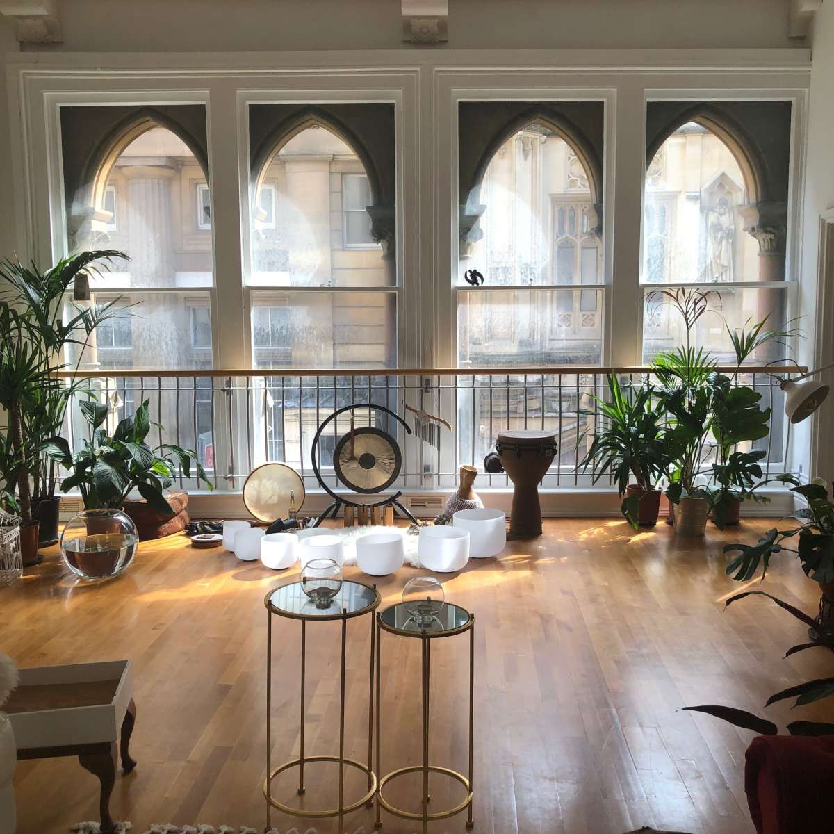 A bright room with large arched windows, featuring various plants, decorative objects, and tables on a wooden floor.