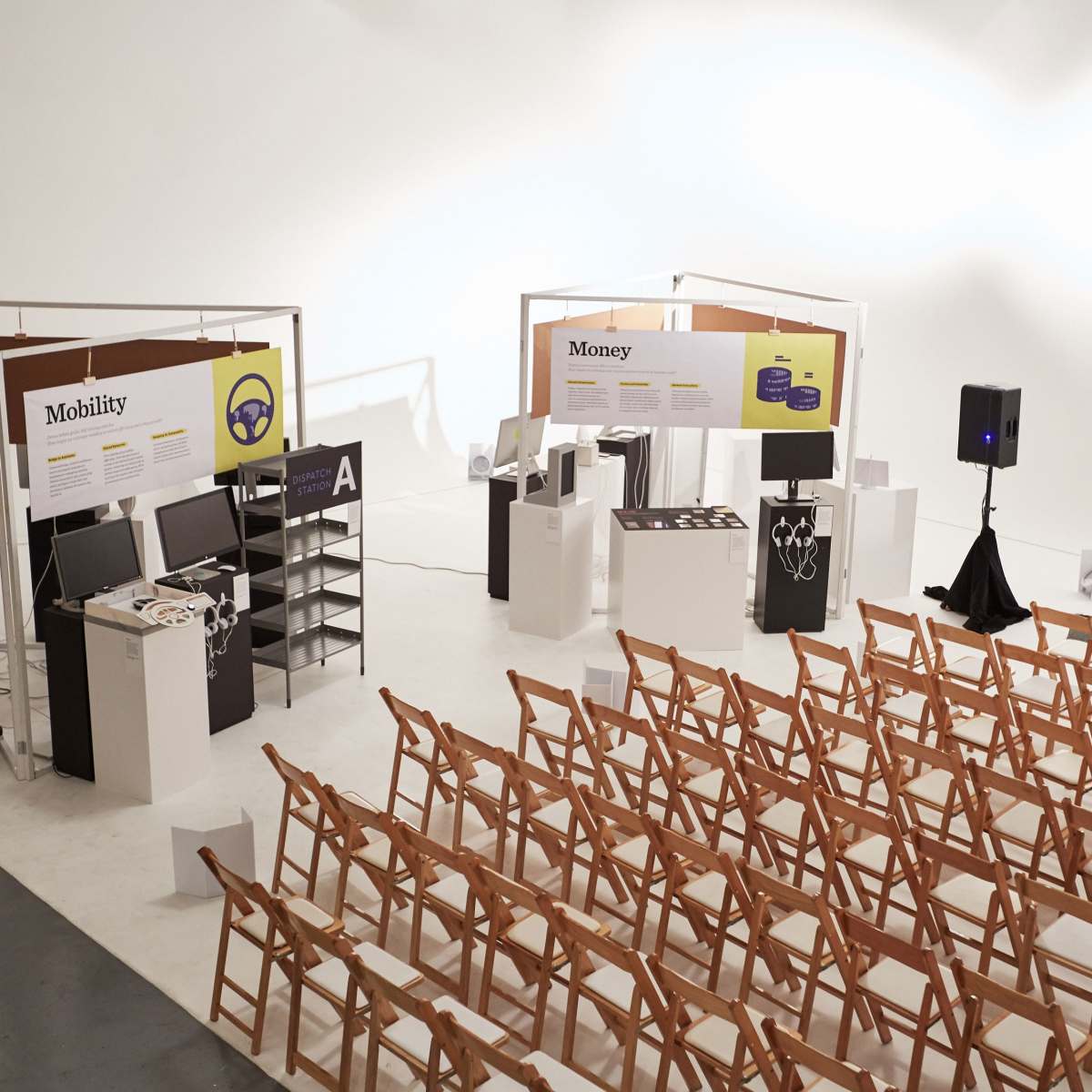 A spacious exhibition setup featuring displays labeled "Mobility" and "Money," with rows of wooden folding chairs arranged in front.