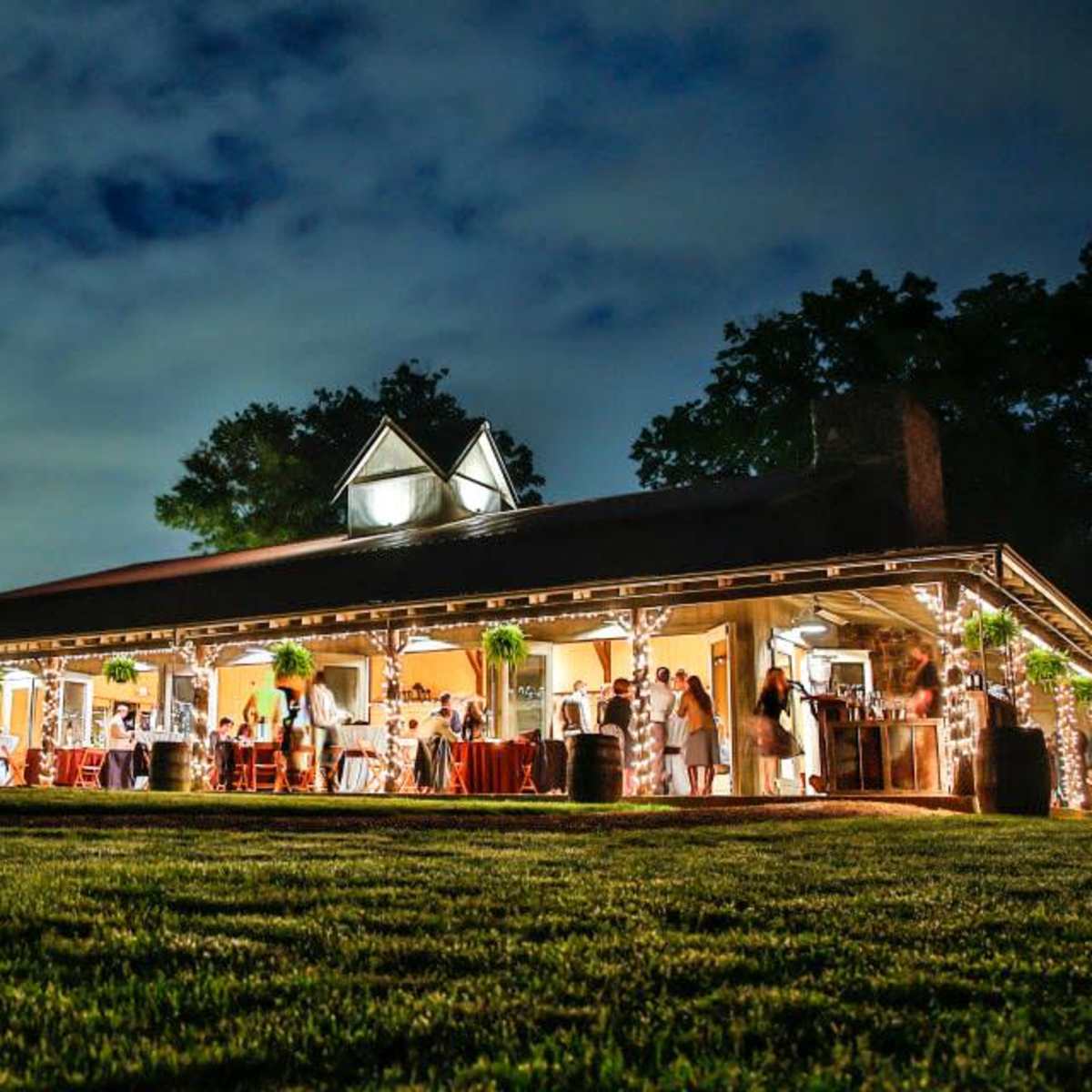 A large, well-lit building with a covered porch hosted a gathering of people in a grassy area under a cloudy sky.