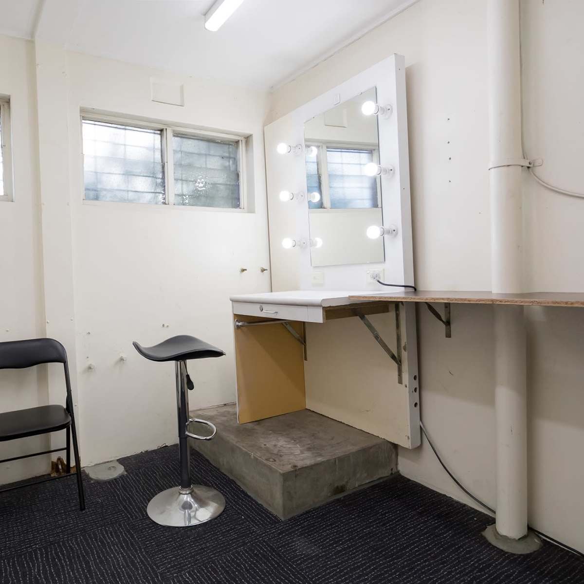 A sparse room with a makeup station featuring illuminated mirrors, a stool, and two collapsible chairs against the wall.