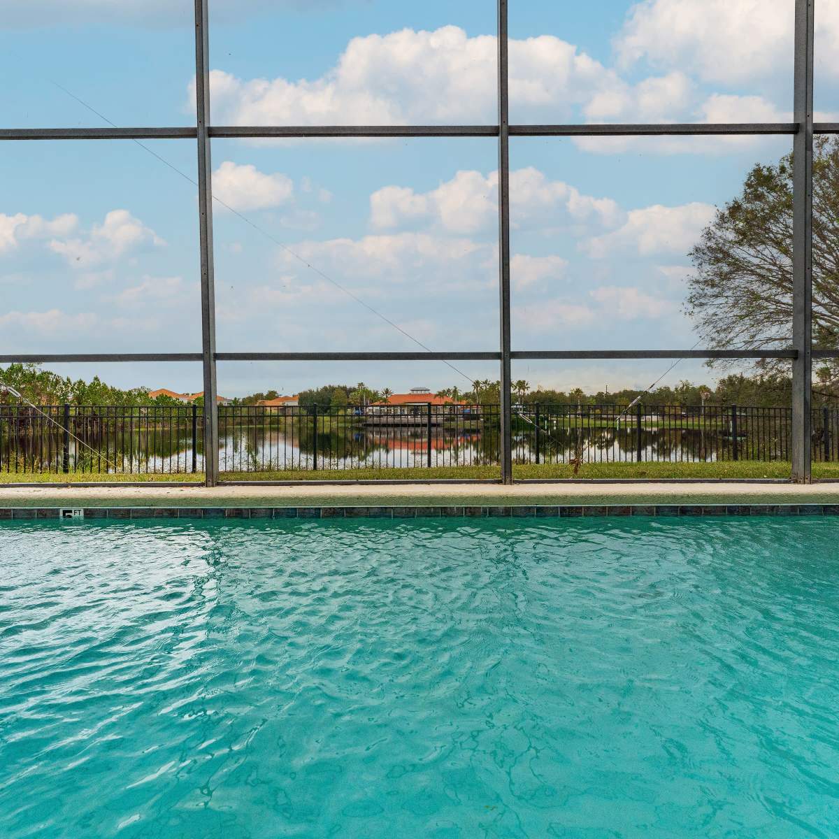 A clear swimming pool is visible through a screened enclosure, with a tranquil view of trees and a lake in the background.