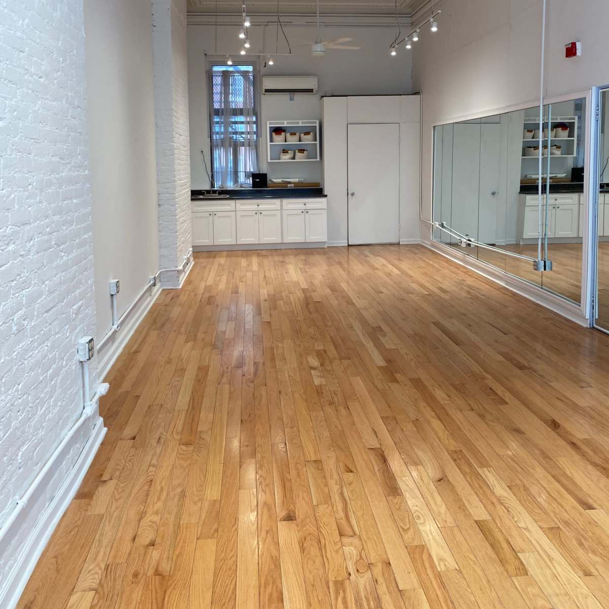 A well-lit, empty room with hardwood flooring, a large mirror on one wall, and a kitchenette area.