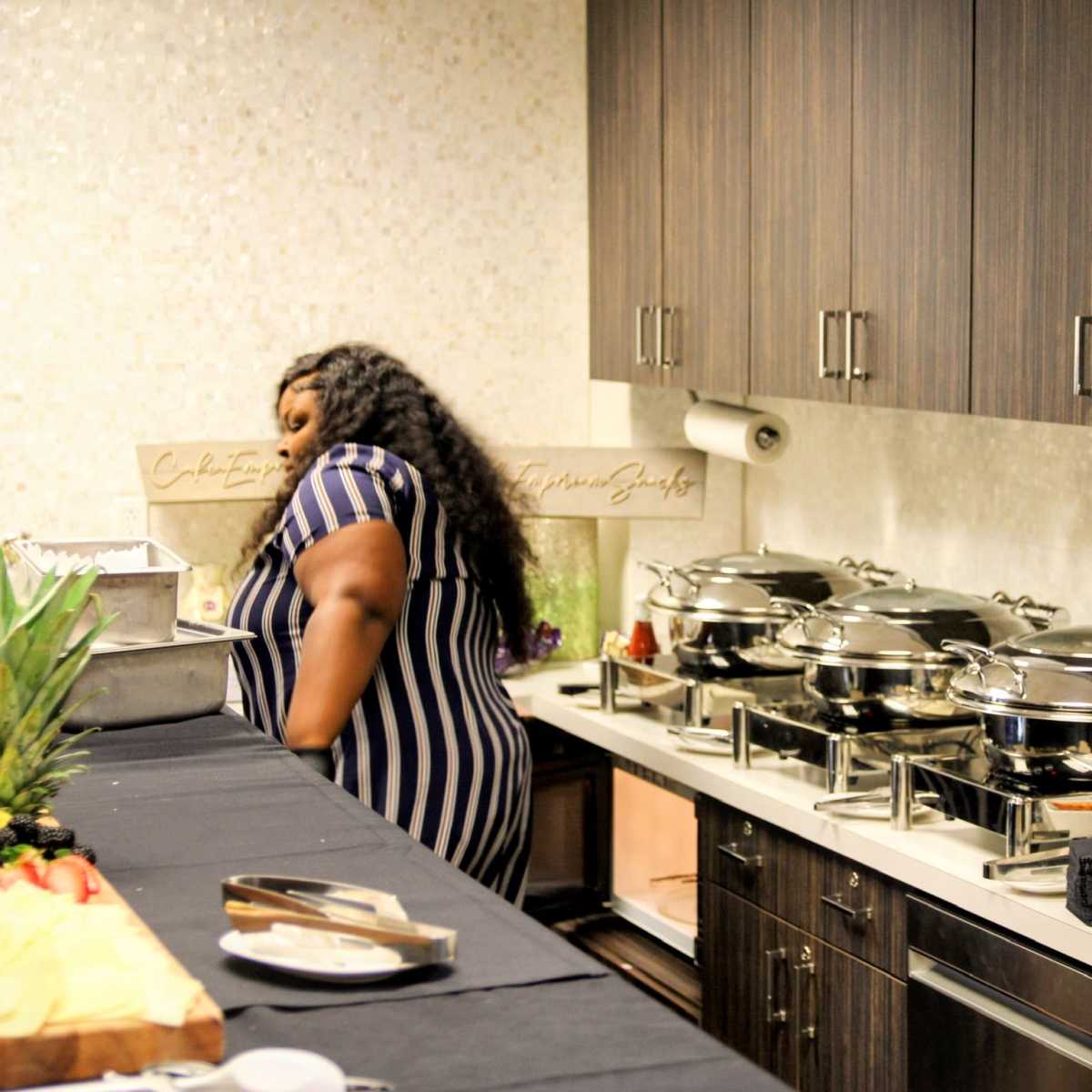 A person in a striped dress stands near a buffet table with three large chafing dishes and stacked plates in a modern kitchen.