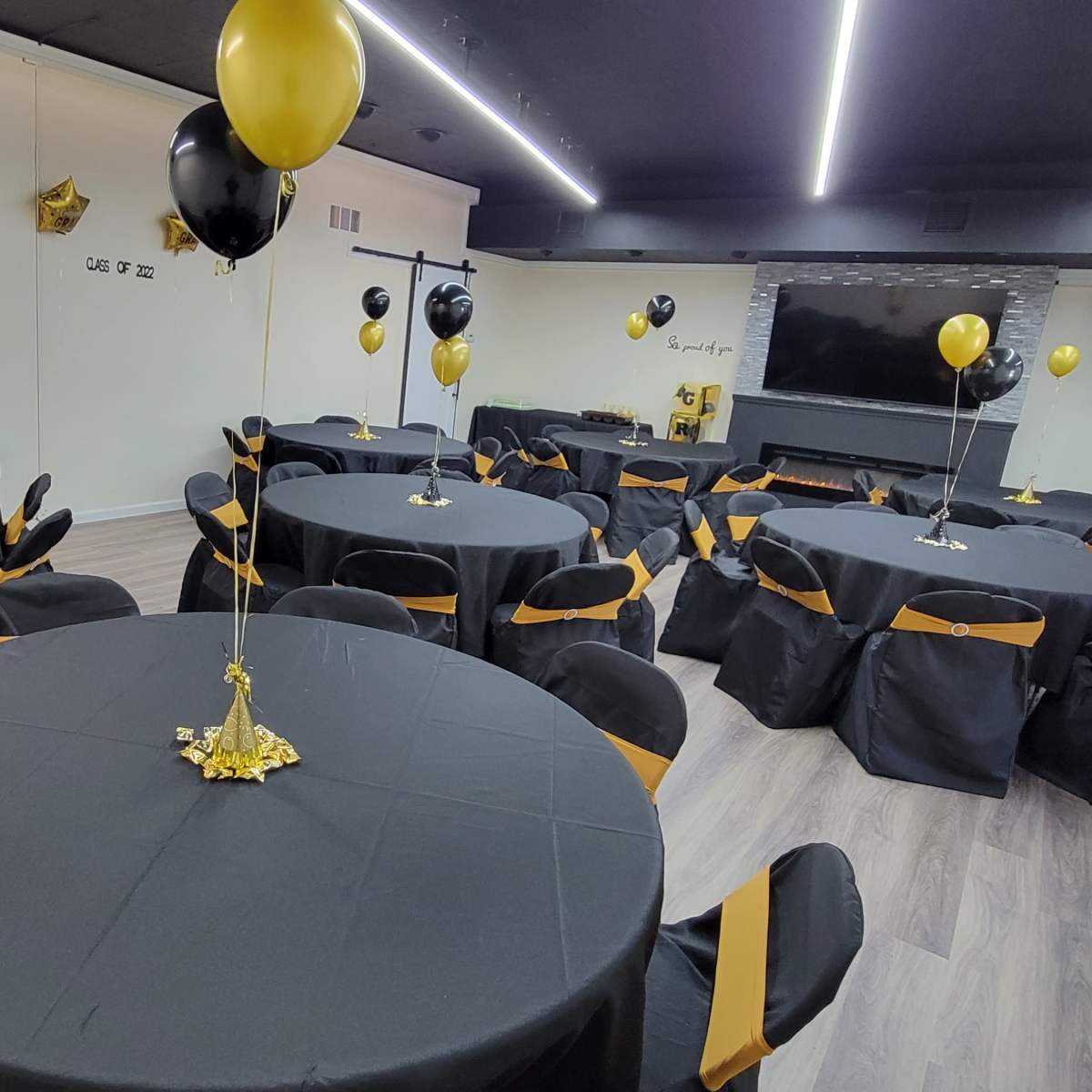 A spacious event room is set up with several round tables covered in black tablecloths, each adorned with gold and black balloons and sashes.