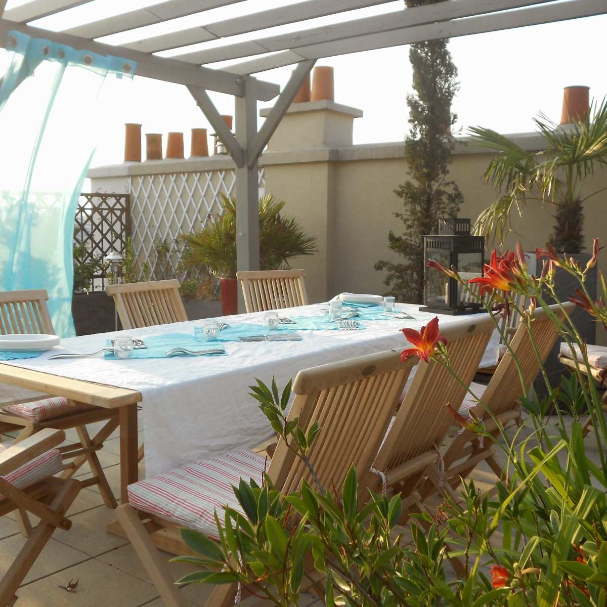 A wooden dining table is set with blue glassware and surrounded by chairs on a terrace adorned with potted plants.
