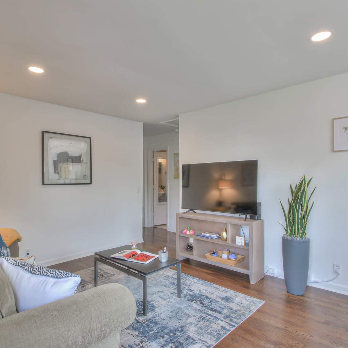 A modern living room features a beige sofa, a coffee table with decorative items, and a television on a light wooden stand, surrounded by minimalist decor.
