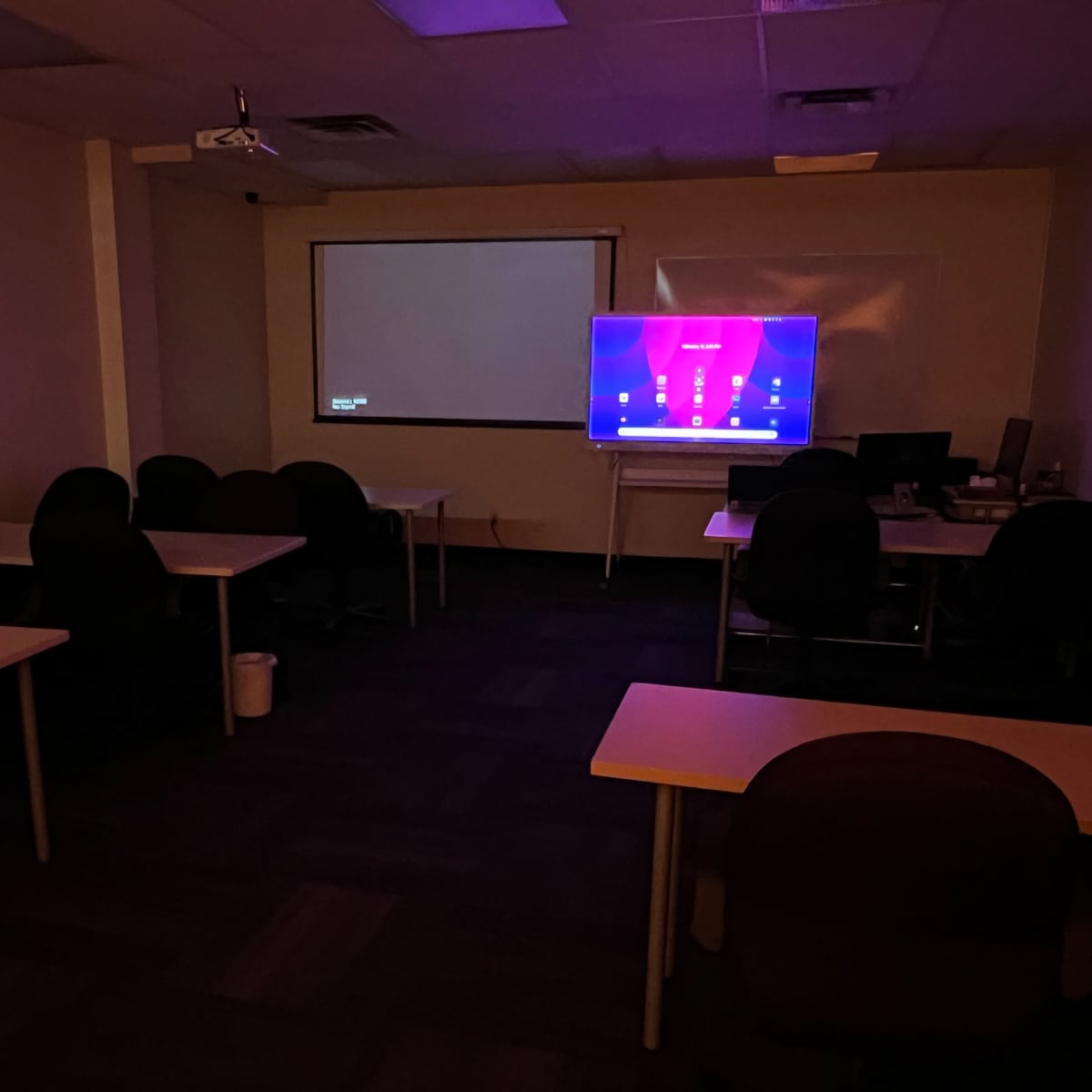 A dimly lit classroom features several empty tables and chairs, with a screen displaying a colorful interface on one wall.