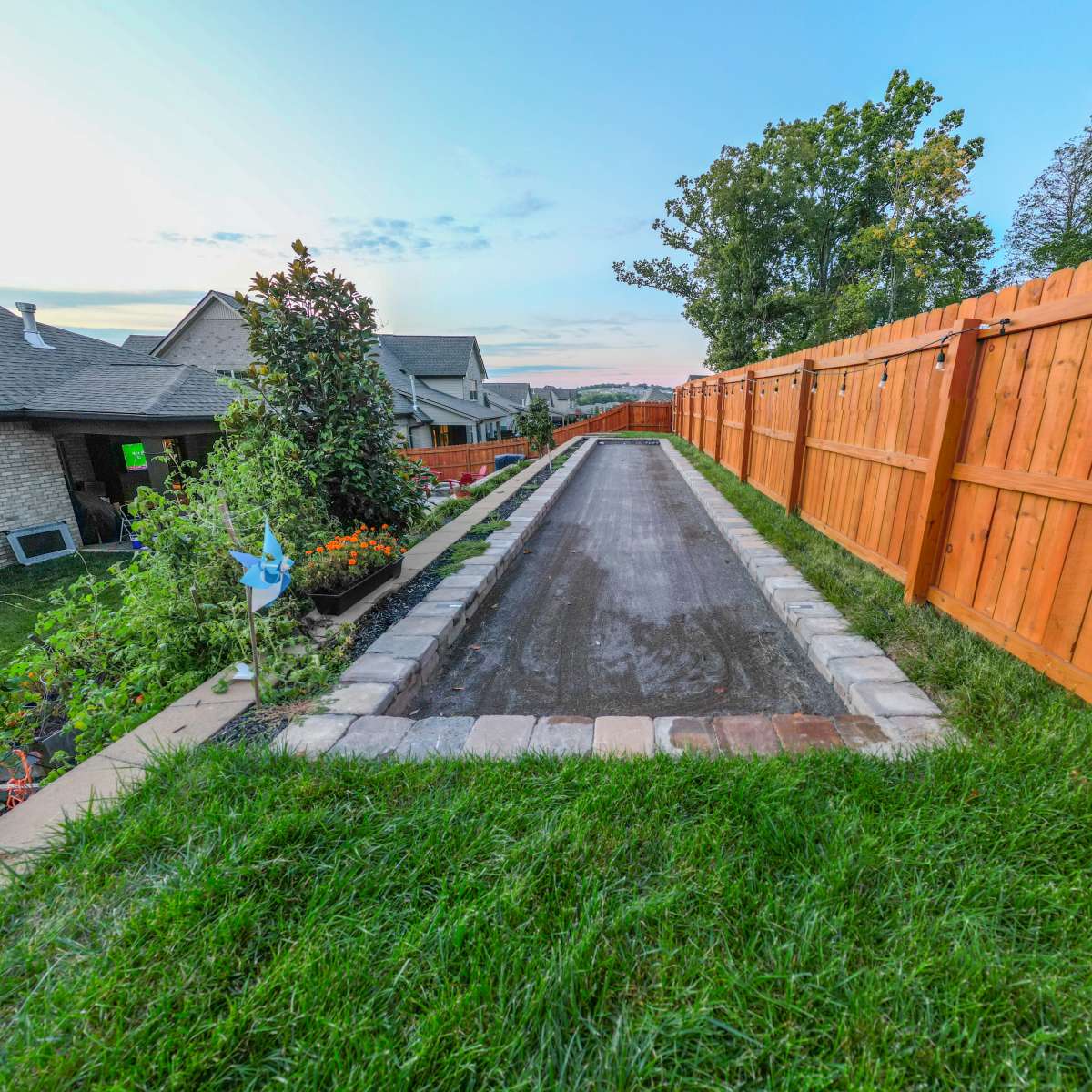 A narrow gravel path bordered by a stone edge, flanked by a tall wooden fence and grassy areas on either side.