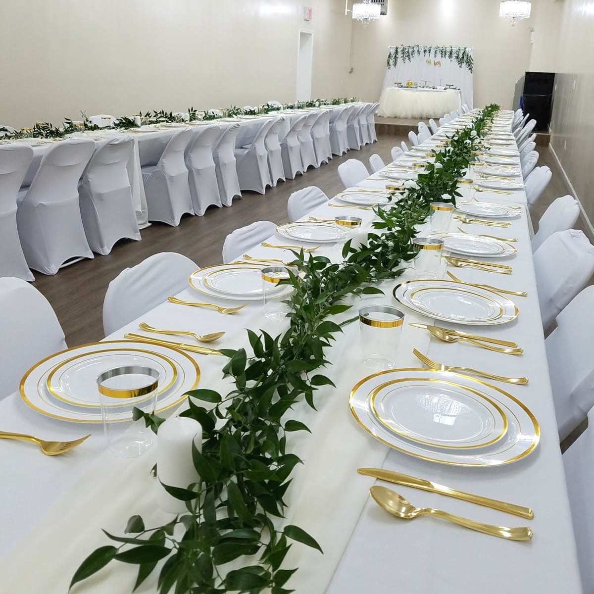 A long banquet table is meticulously set with white tablecloths, gold-accented plates, and a centerpiece of greenery, arranged in a spacious, well-lit dining area featuring chandeliers.
