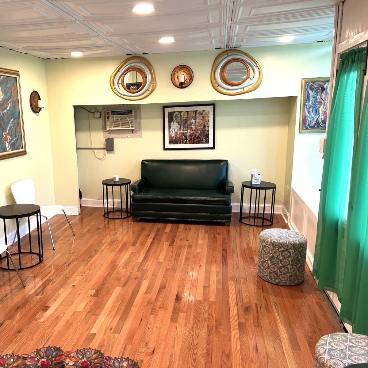 The room features a green sofa, two white chairs, small black tables, and several framed artworks on the walls, with wooden flooring and a green curtain.
