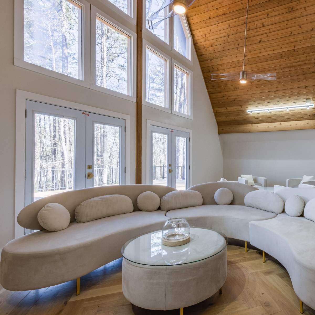 A modern living room featuring a curved beige sofa, a glass coffee table, and large windows that overlook a wooded area.