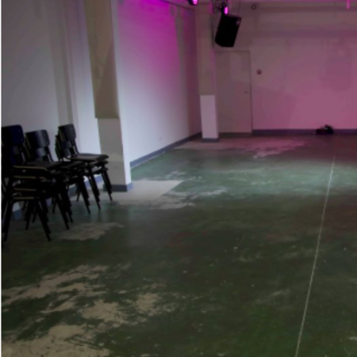 An empty, sparsely furnished room with green flooring, a few stacked chairs against one wall, and ambient lighting.