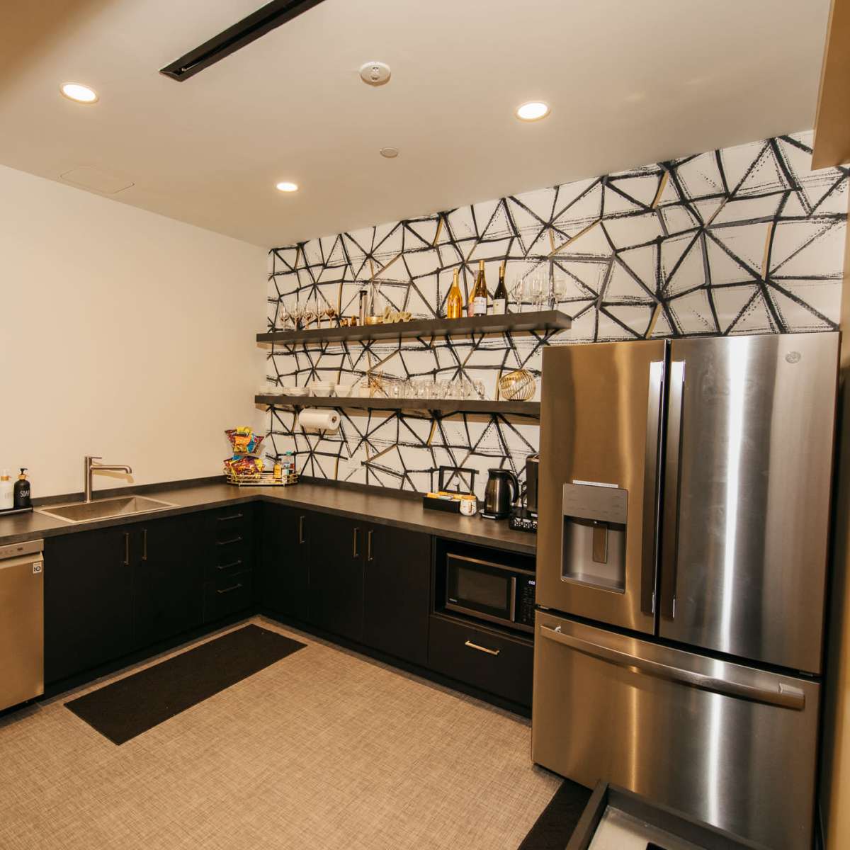 A modern kitchenette featuring dark cabinetry, a stainless steel refrigerator, and geometric-patterned wall shelves with various kitchen items.