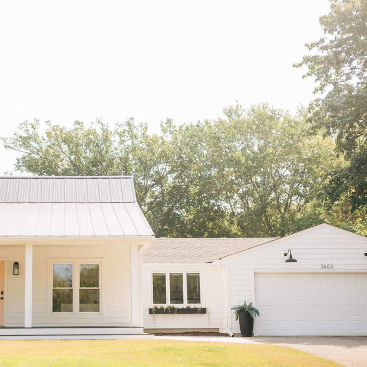 A white house with a metal roof, large windows, and an attached garage, set against a backdrop of trees.