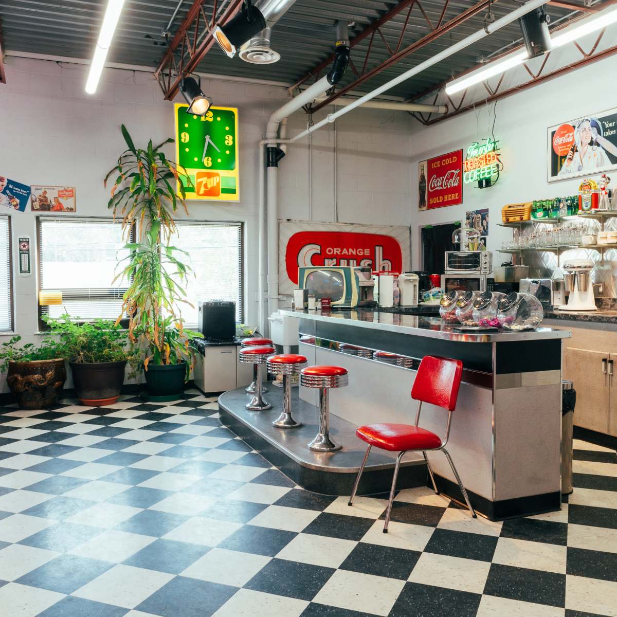A retro diner with red chairs, a checkered floor, and neon signs, featuring a counter with stools and various vintage decorations on the walls.