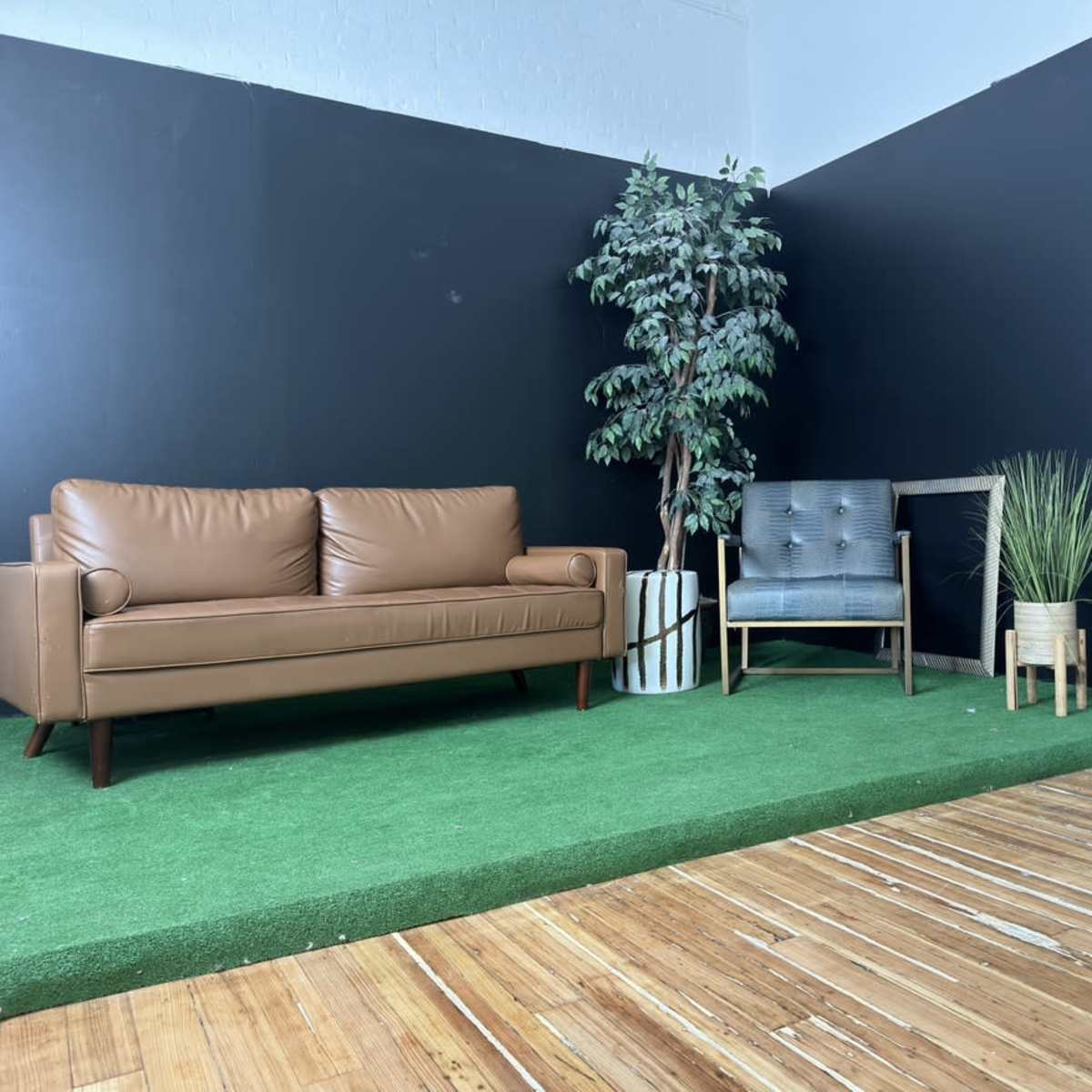 A small interior space with a brown couch, a gray chair, decorative plants, and a wooden floor.