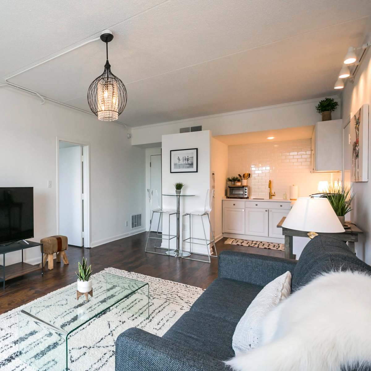 A modern living room features a gray sofa, a glass coffee table, a television on a stand, and a kitchen area visible in the background.