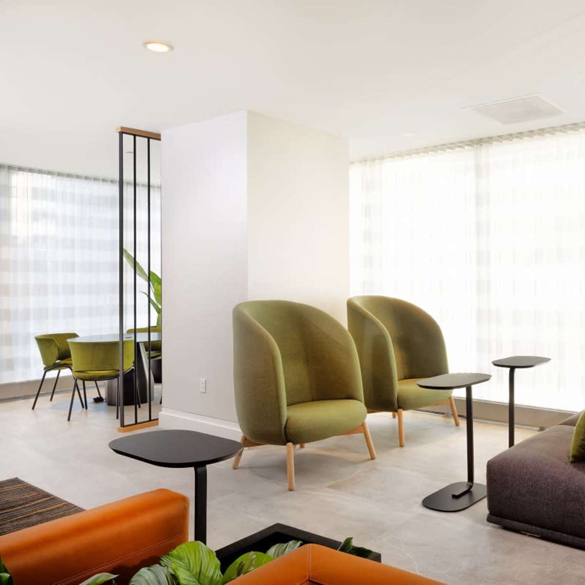 A modern lounge area with green and gray furniture, small tables, and large windows allowing natural light to enter.