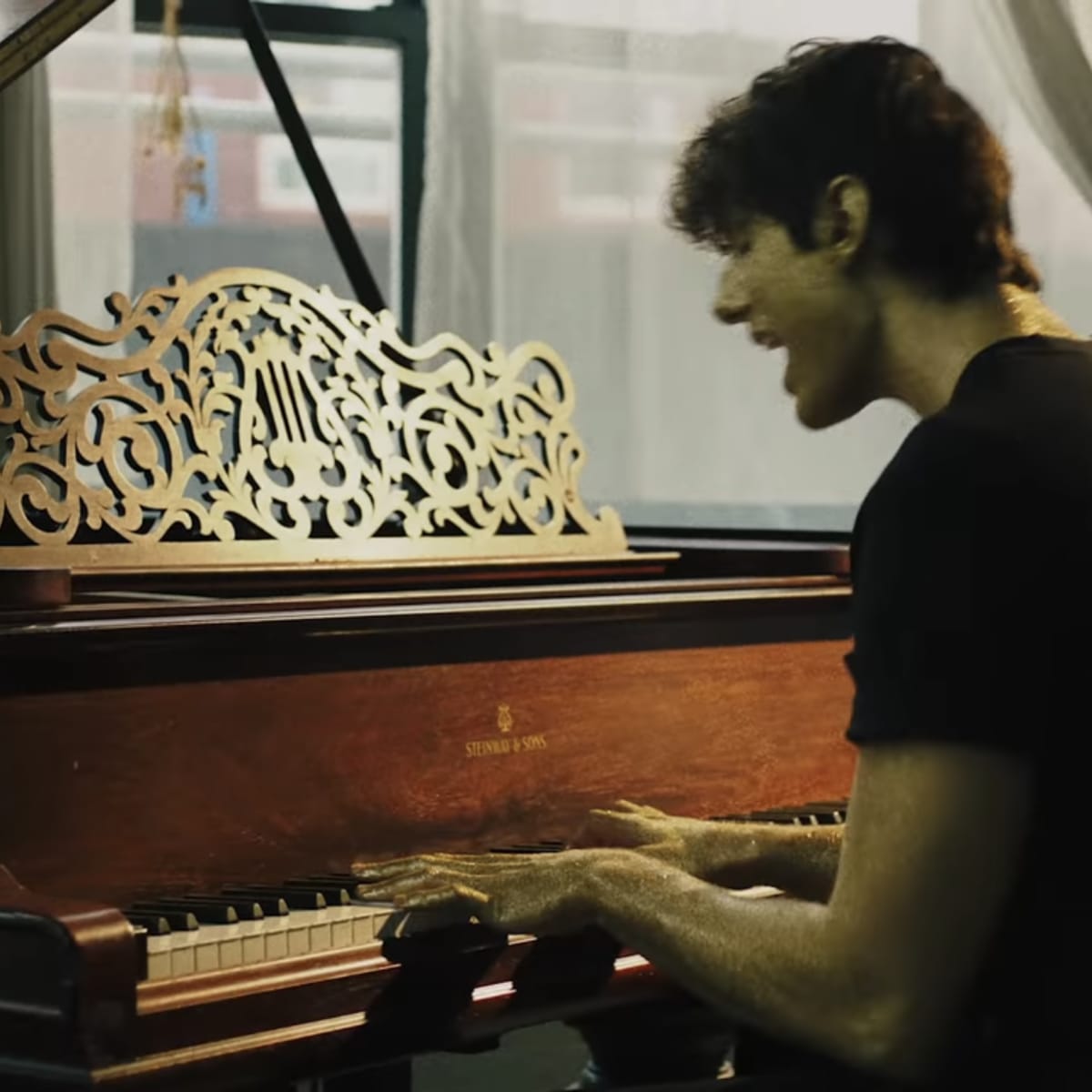 A young man is playing a wooden piano in a well-lit room with large windows.