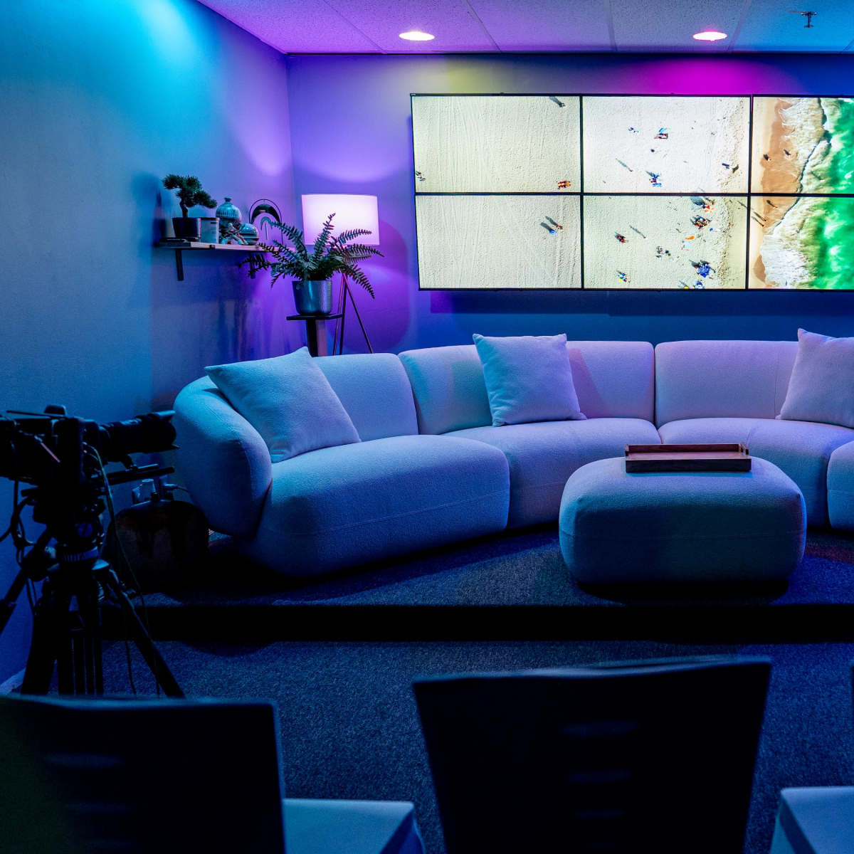 A modern lounge area features a white sectional sofa, a central wooden coffee table, and multiple cameras aimed at a large screen displaying a beach scene.