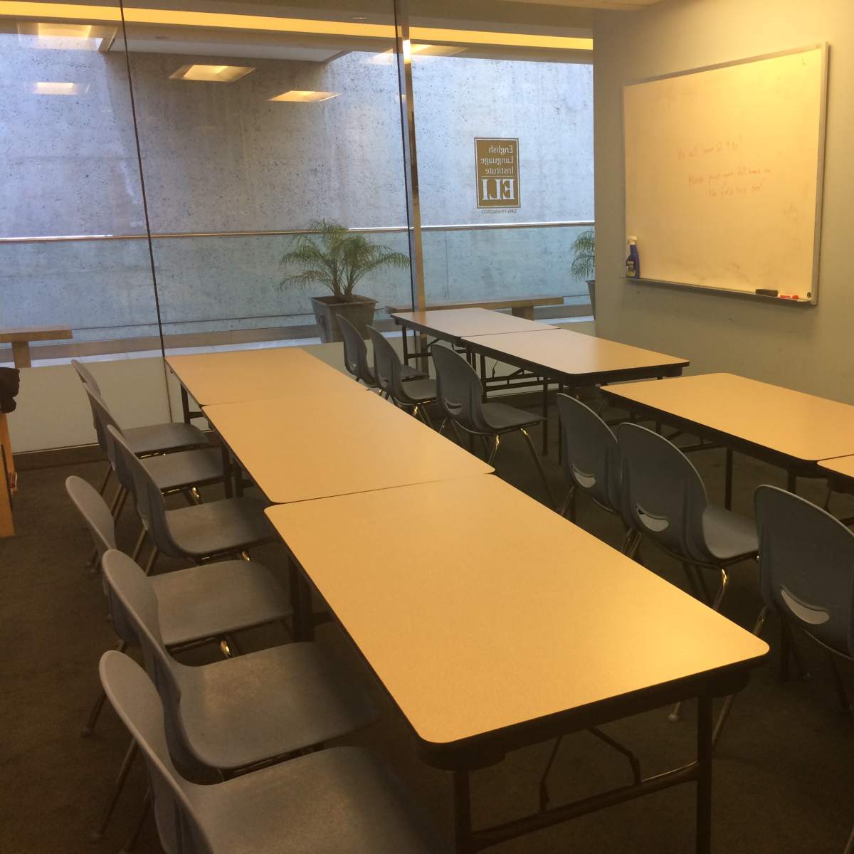 A classroom with several tables arranged in two rows, each accompanied by blue chairs, and a whiteboard and TV mounted on the wall.