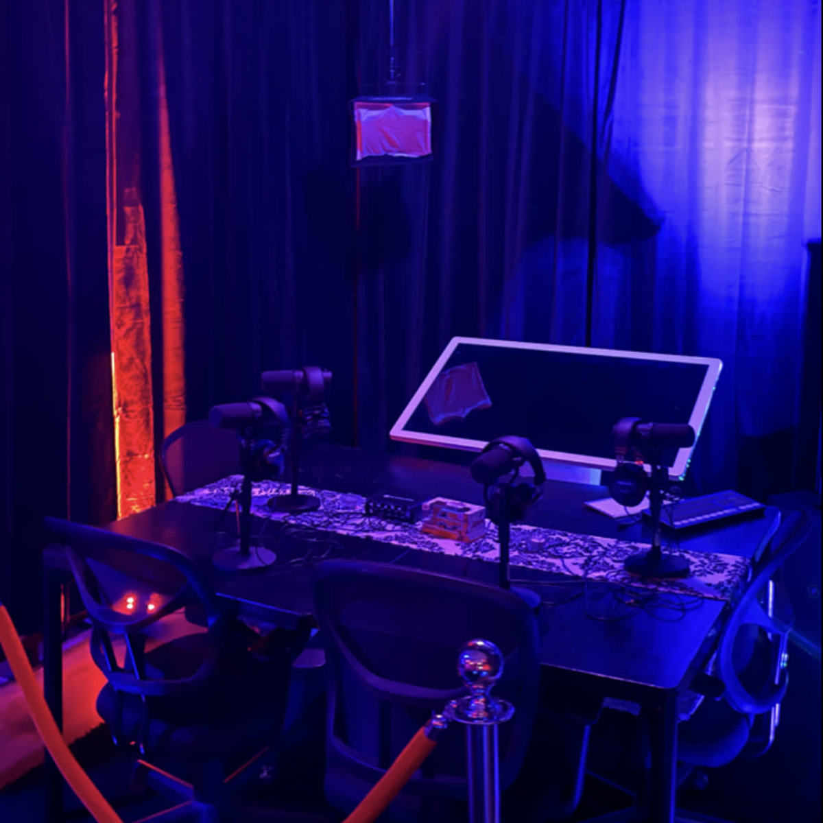 A dimly lit room with blue and purple lighting, featuring a table with microphones and a large monitor surrounded by dark curtains.
