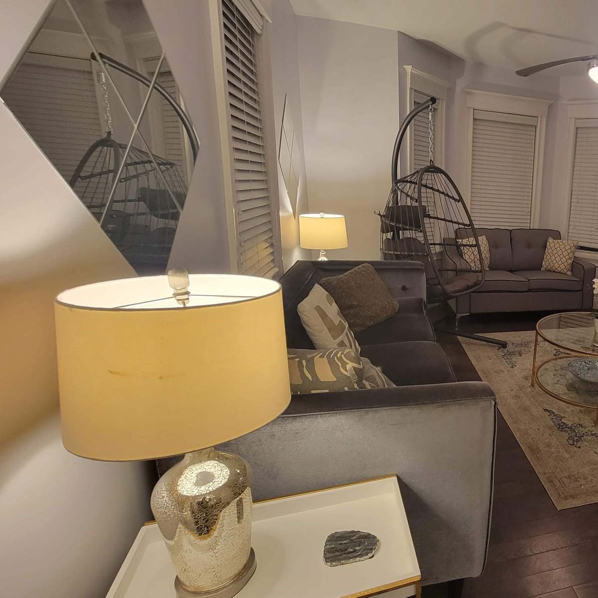 A living room with two gray sofas, a glass coffee table, and a spiral staircase, all illuminated by a table lamp and overhead light.