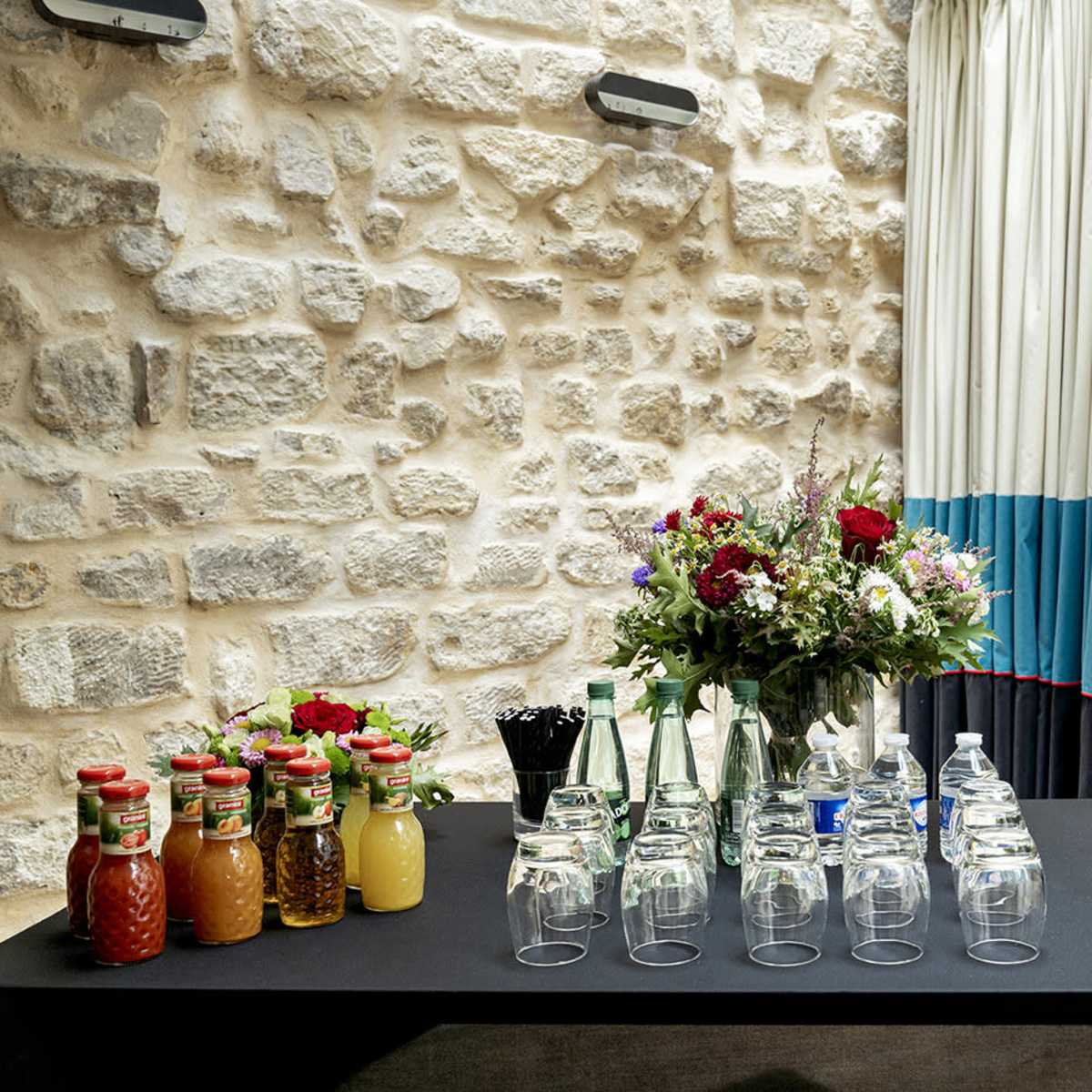 A table is set against a stone wall, displaying various bottles of juices, sodas, and glasses, alongside a floral arrangement.