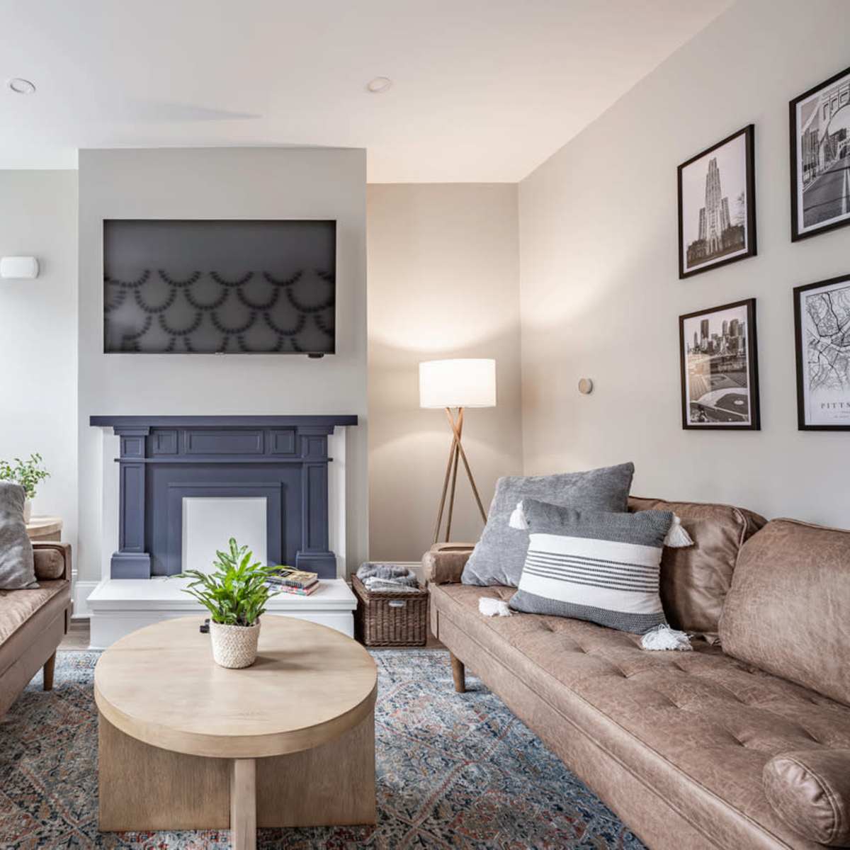 A cozy living room features two brown sofas facing a circular wooden coffee table, a blue fireplace with a TV above, and light gray curtains.