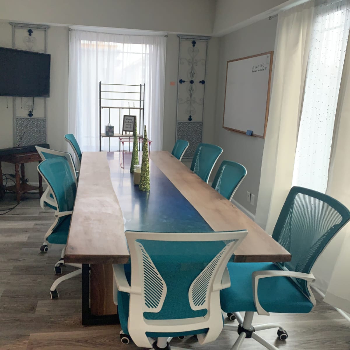 A conference room with a long wooden table surrounded by turquoise chairs, a television mounted on the wall, and a whiteboard in the background.