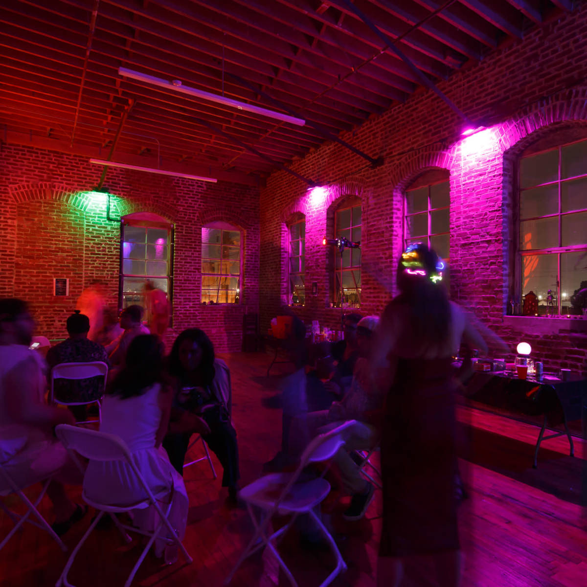 A vibrant gathering takes place in a brick-walled room lit with colorful lights, where people are seated and mingling.