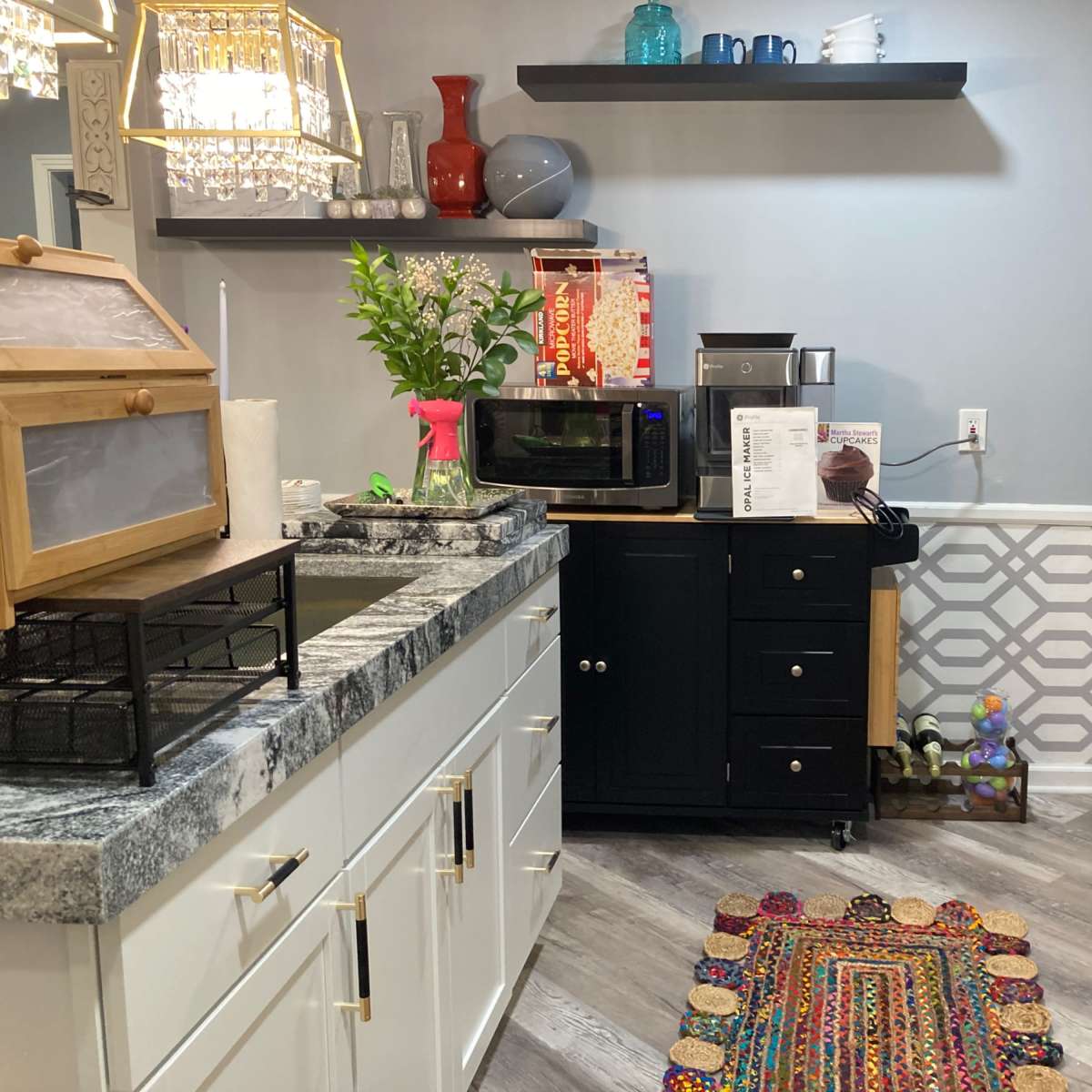 A kitchen area with a black microwave, a small black cabinet, and decorative items on shelves, along with a colorful rug on the floor.
