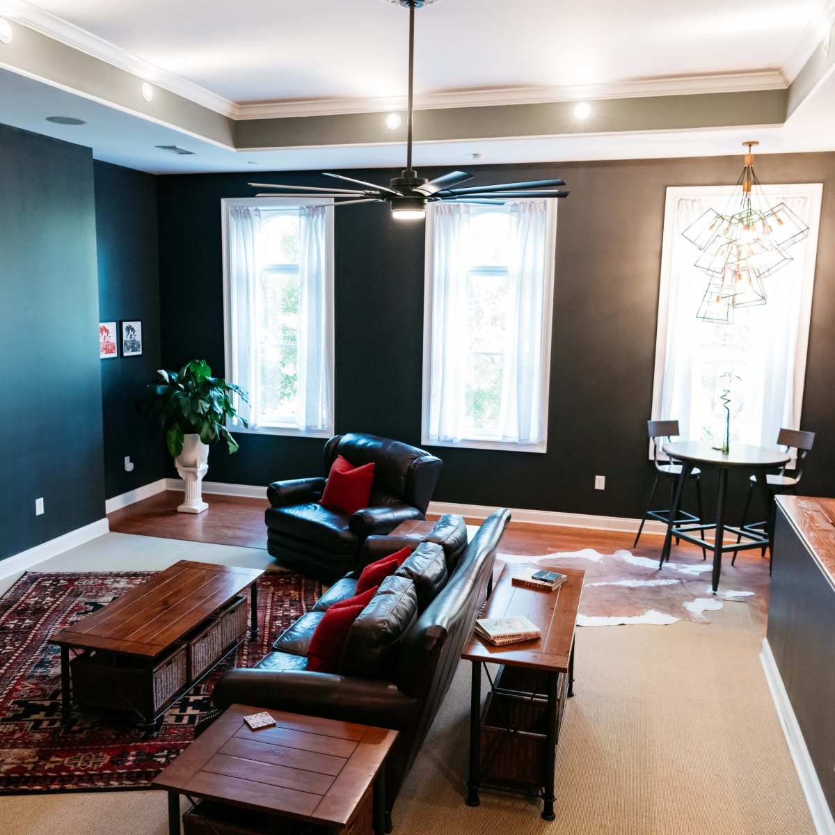 A spacious living room features a leather couch, a coffee table, and large windows adorned with sheer curtains, illuminated by overhead lights.