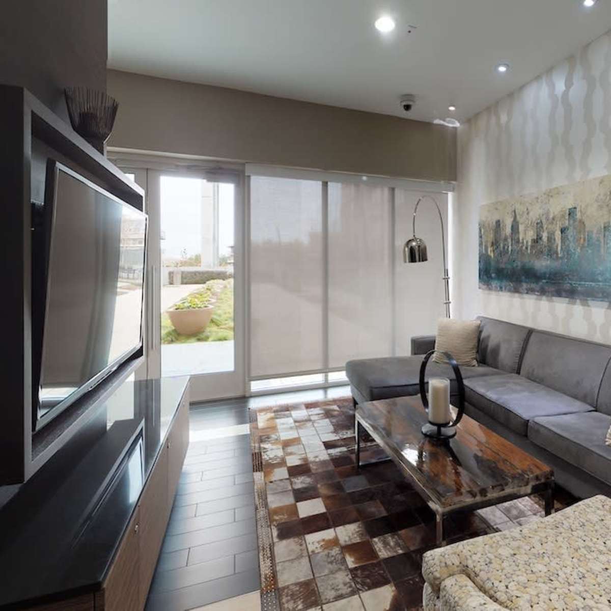 A modern living room features a gray sofa, a wooden coffee table, and large windows with blinds, overlooking a landscaped area.