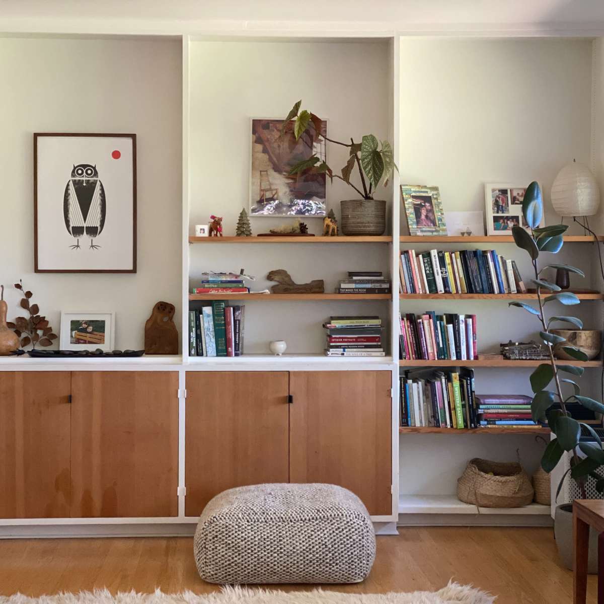 A modern living room with a wooden shelving unit filled with books and decorative items, accompanied by a round pouf and a leafy plant in the foreground.