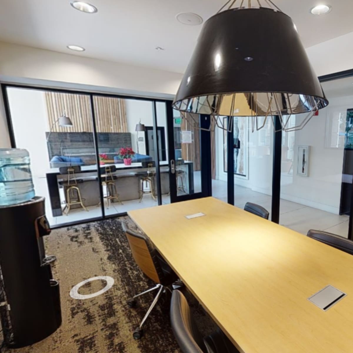 A modern conference room with a wooden table, black chairs, and glass walls, leading to a hallway and another room.
